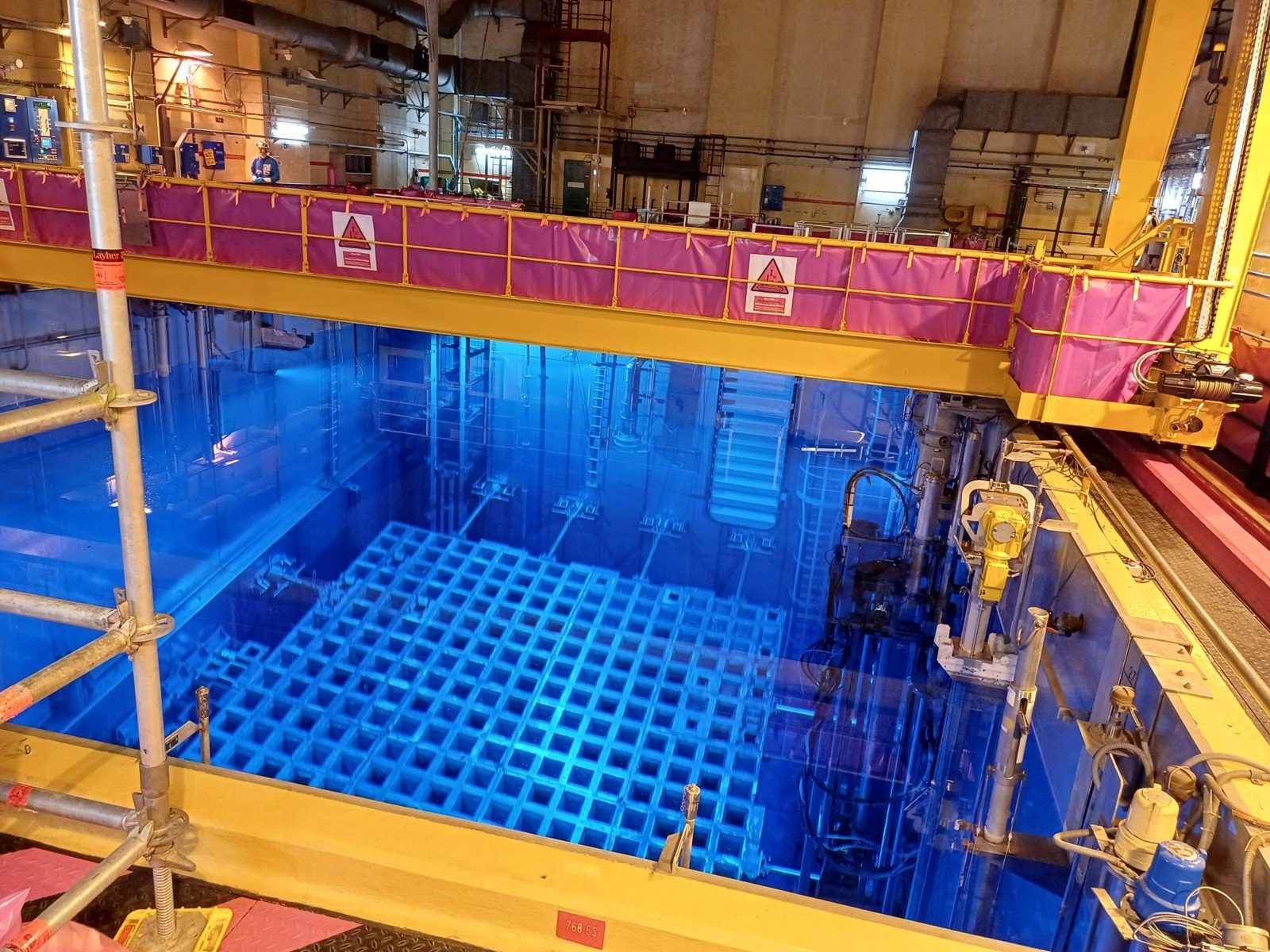 Piscine du bâtiment réacteur d’une centrale nucléaire pendant un arrêt de tranche : eau d’un bleu intense, râteliers de stockage des assemblages visibles au fond, pont de manutention jaune bordé de bâches de protection roses portant des panneaux de zone contrôlée, échafaudage au premier plan et un intervenant en tenue au loin sur la passerelle. Photo prise lors d’une intervention PCR Group